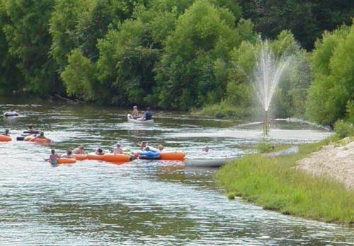 People in colorful inflatable boats float on a river, surrounded by greenery and a fountain in the background.