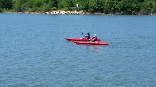 Two people in red kayaks paddle on a calm lake surrounded by green trees.