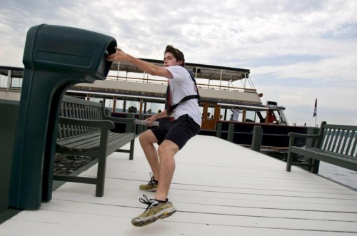 A young man in a harness pulls on a dockside post near a boat, with cloudy skies in the background.