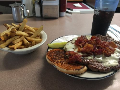 A plate with a cheeseburger topped with bacon and a pickle, served with a side of fries and a drink.