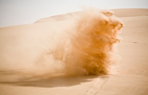 A cloud of sand billows up from a desert dune, creating a dynamic and textured scene in warm tones.