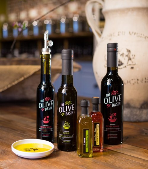 A selection of olive oils and vinegars displayed on a wooden table, with a small dish and a decorative jug in the background.