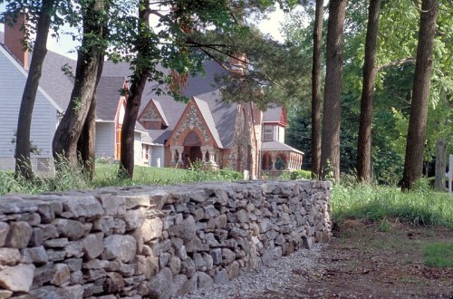A stone wall in the foreground with a large, elegant house and trees in the background on a sunny day.