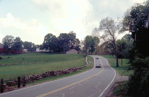 A winding road leads through a rural landscape with trees and a house in the background under a cloudy sky.