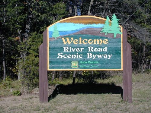 Welcome sign for River Road Scenic Byway, featuring trees and a landscape design, located in Huron-Manistee National Forest.