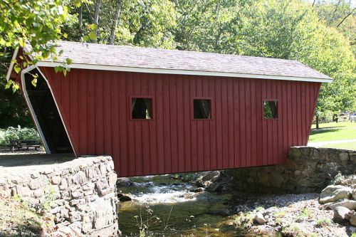 A red covered bridge spans a small stream, surrounded by green trees and a grassy area.