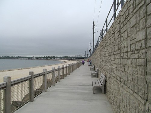 A concrete walkway along a beach, with benches and a stone wall, under a cloudy sky. People stroll in the distance.