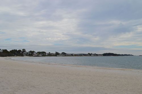 A serene beach scene with soft sand, calm water, and a distant shoreline under a cloudy sky.