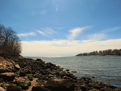 A serene coastal view with rocky shorelines, calm water, and a clear blue sky dotted with wispy clouds.