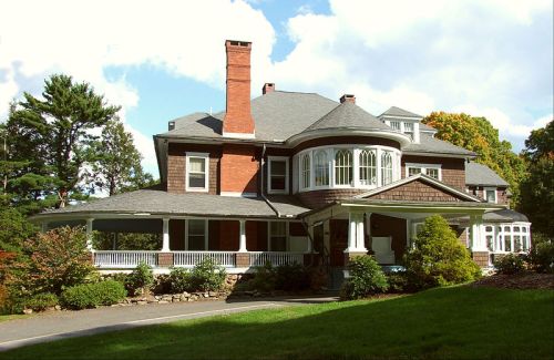 A large, historic house with a wraparound porch, multiple gables, and a prominent chimney, surrounded by greenery.