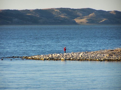 A person in a red shirt stands on a rocky shore by a calm lake, with hills in the background under a clear sky.