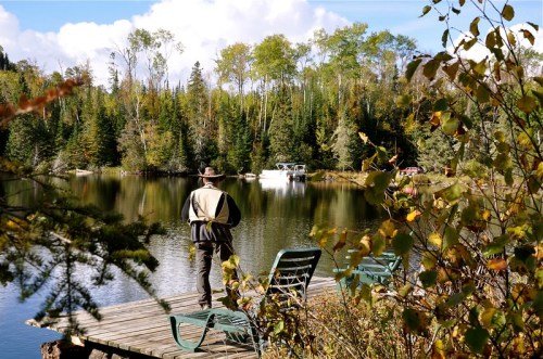 A person walks along a dock by a calm lake surrounded by trees and autumn foliage.