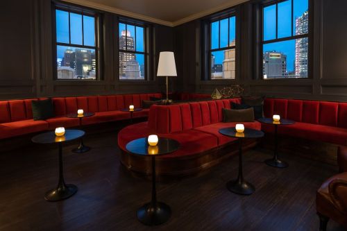 A stylish lounge with red seating, small tables, and soft lighting, featuring city views through large windows at dusk.
