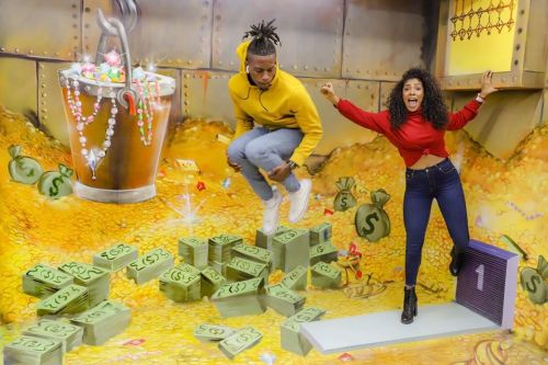 Two people pose in a colorful room filled with oversized money bags and gold coins, one jumping and the other excitedly posing.