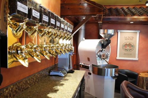 A coffee shop interior featuring a row of glass containers for beans and a coffee roaster against a warm-colored wall.