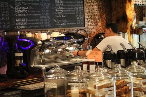 A barista prepares coffee at a café, with a menu board and jars of ingredients in the foreground.