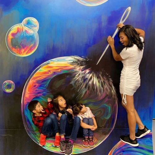 A woman interacts with children inside a colorful, painted bubble, creating a playful and imaginative scene.