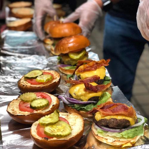 A row of freshly assembled burgers with lettuce, tomato, pickles, cheese, and bacon on toasted buns.