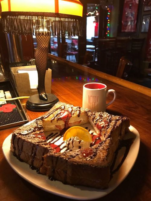 A plate of French toast topped with whipped cream, chocolate drizzle, and a lemon slice, next to a festive mug and lamp.