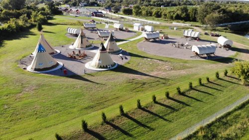 Aerial view of a campsite featuring teepees and covered wagons set in a grassy area with trees in the background.