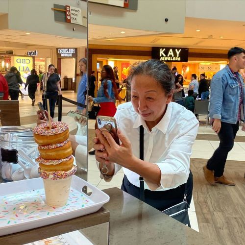 A woman takes a photo of a tall stack of donuts with sprinkles and a milkshake in a busy mall setting.