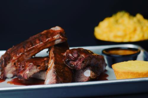 A plate of barbecue ribs with sauce, a slice of cornbread, and a side of macaroni and cheese in the background.