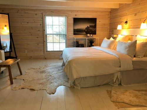 Cozy bedroom with a large bed, wooden walls, a mirror, and soft lighting, featuring a rug and a window.