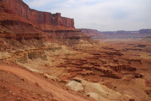 A vast, rugged canyon landscape with layered red rock formations and a winding dirt path.