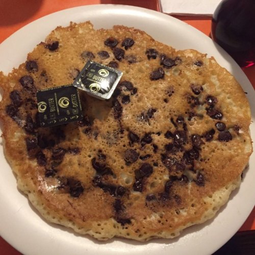 A large, golden-brown pancake topped with chocolate chips and two butter packets on a white plate.