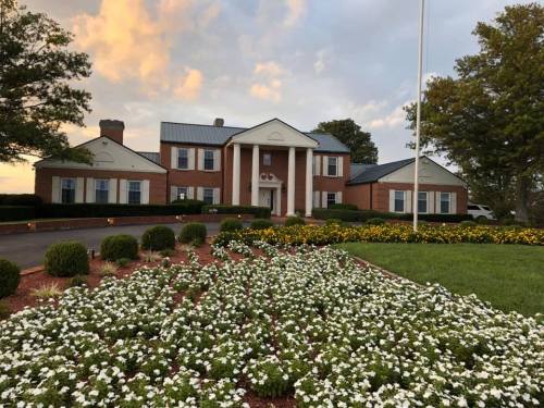 A large brick house with a white porch, surrounded by colorful flower beds and green lawns under a cloudy sky.