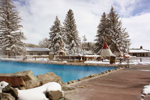 A serene winter scene featuring a blue pool surrounded by snow-covered trees and tipis.