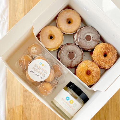 A box of assorted donuts, including glazed, chocolate, and sugar-coated, alongside banana pudding donut holes.