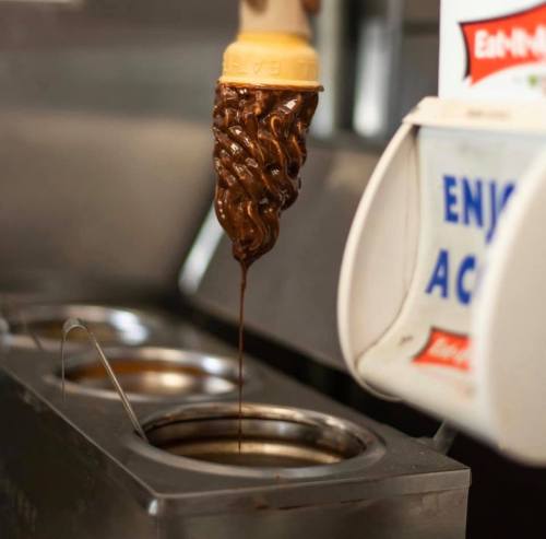 A chocolate ice cream cone being filled with thick chocolate syrup from a dispenser.