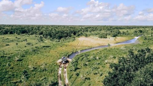 Aerial view of a winding river through lush green fields and trees, with a vehicle on a dirt path.