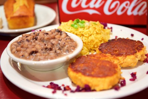 A plate featuring black-eyed peas, yellow rice, and two golden corn fritters, with a Coca-Cola backdrop.