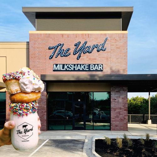 A hand holds a colorful milkshake topped with donuts and sprinkles in front of The Yard Milkshake Bar.
