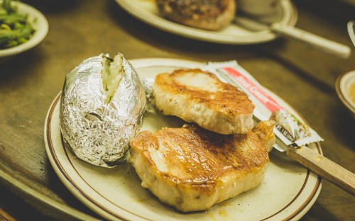 A plate with two golden-brown pork chops, a baked potato wrapped in foil, and a butter packet.