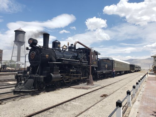 A vintage steam locomotive with passenger cars, parked on tracks under a blue sky with clouds.