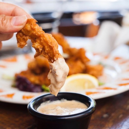 A hand dips a crispy fried shrimp into a small bowl of creamy sauce, with more shrimp on a plate in the background.