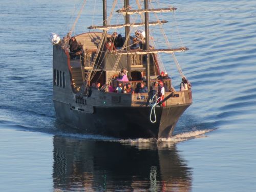 A wooden pirate ship with passengers sails on calm waters, creating ripples in the reflection.