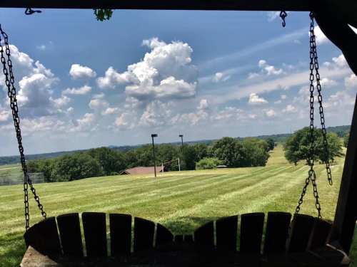 A scenic view from a swing, overlooking a green field and distant hills under a blue sky with fluffy clouds.