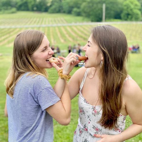Two women playfully feeding each other snacks in a vineyard, with lush green fields in the background.