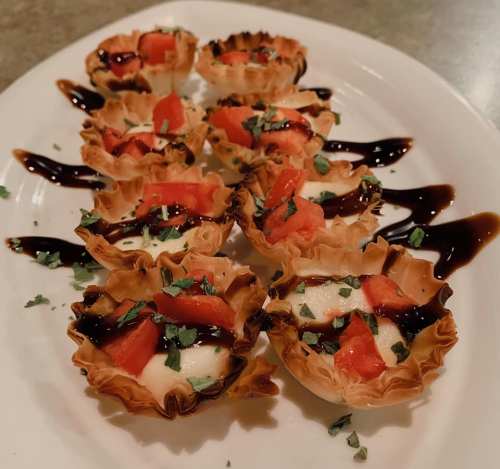 A platter of mini phyllo cups filled with cheese, topped with diced tomatoes and drizzled with balsamic glaze.