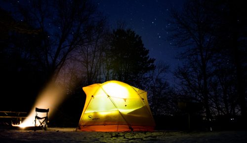 A glowing tent under a starry night sky, with a camp chair nearby and a beam of light illuminating the scene.