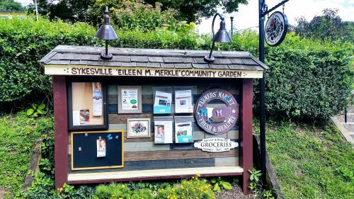 Signboard for Sykesville Eileen M. Merkle Community Garden, featuring community notices and grocery information.