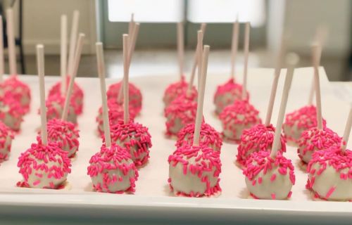 A tray of cake pops coated in white chocolate and topped with pink sprinkles, each on a stick.