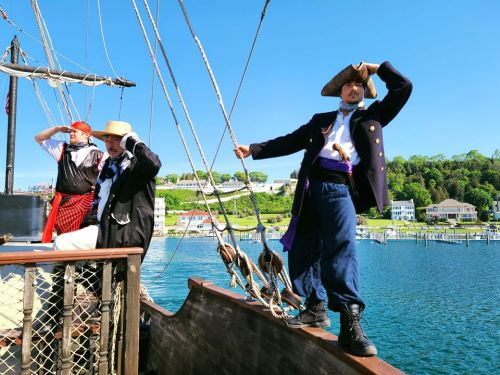Two men in pirate costumes stand on a ship, one saluting and the other looking out at the water, with a scenic background.