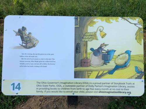 A sign featuring a storybook illustration of a mouse and a child reading in a peaceful outdoor setting.