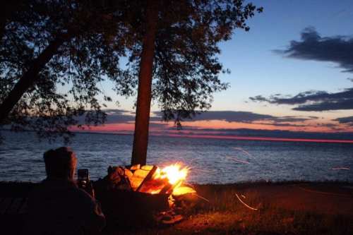 A person sits by a campfire at dusk, with a lake and colorful sunset in the background, surrounded by trees.