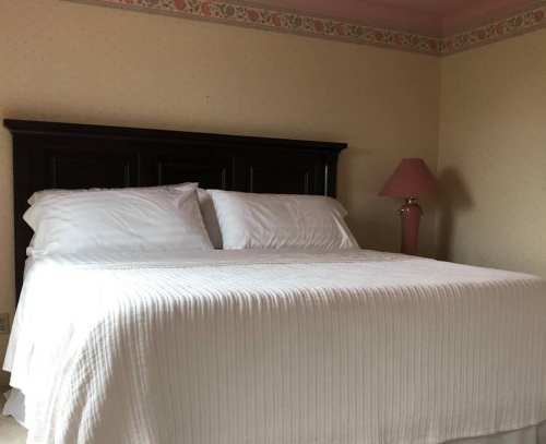 A neatly made bed with white linens in a softly lit room featuring a pink lamp and patterned wallpaper.
